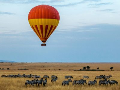 A vast herd of wildebeest crossing the Mara River in the Maasai Mara National Reserve, Kenya, with zebras and other animals in the background, and a hot air balloon floating in the sky above.