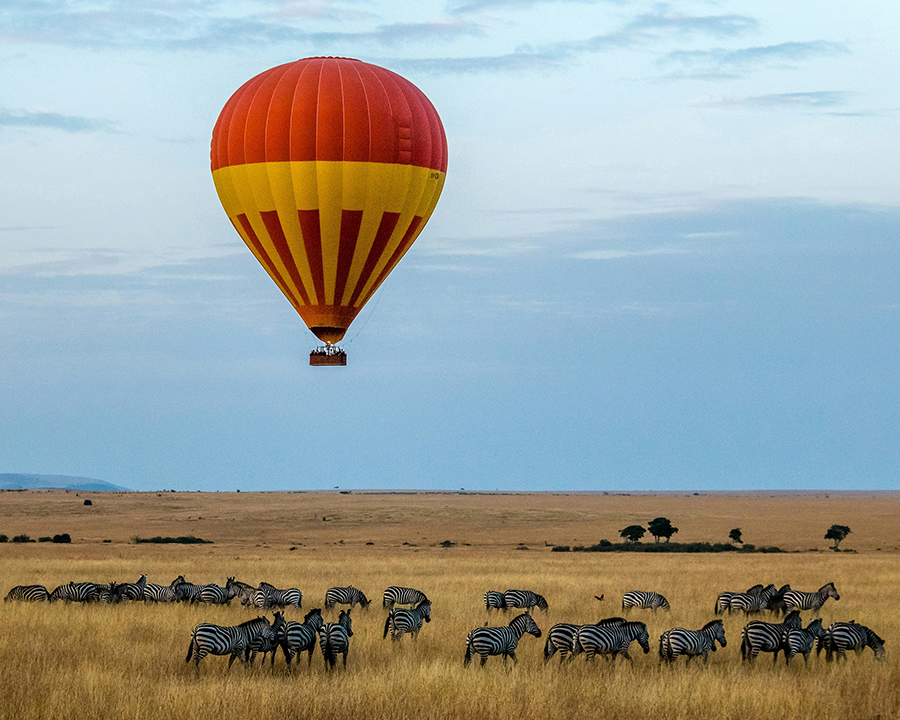 A vast herd of wildebeest crossing the Mara River in the Maasai Mara National Reserve, Kenya, with zebras and other animals in the background, and a hot air balloon floating in the sky above.