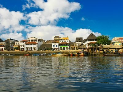 A traditional dhow sailing boat with white sails gliding through the turquoise waters of the Indian Ocean, with the historic town of Lamu in the background.