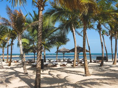 A pristine white sand beach fringed by palm trees and turquoise waters on the Kenyan coast, with a traditional dhow sailing boat in the distance.